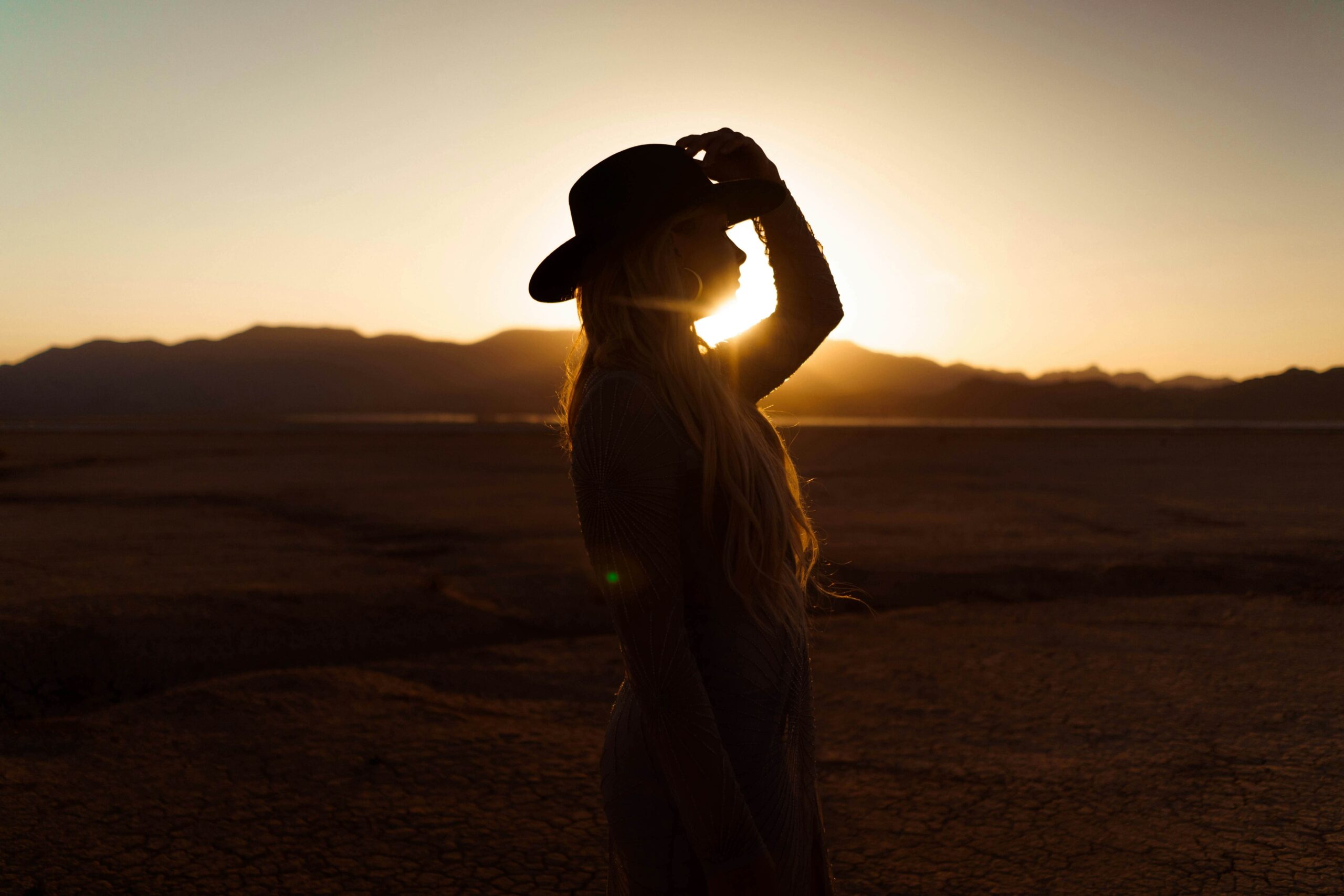 Silhouette of a person wearing a hat stands on dry, cracked ground at sunset with mountains in the background, evoking the signature style of brand-showit-website-designer Sweet Pea Ink Creative.